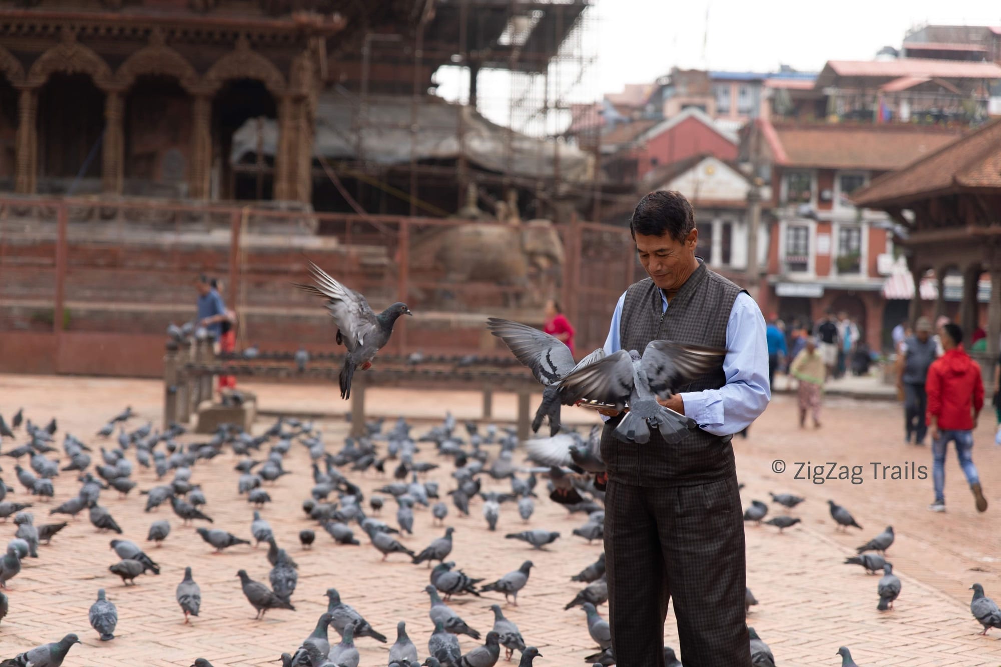 A man smiling while feeding pigeons in front of Krishna Temple in Patan Darbar Square.